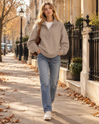 Beige oversized henley sweatshirt with snap button collar worn with straight-leg jeans and white trainers on a tree-lined pavement.