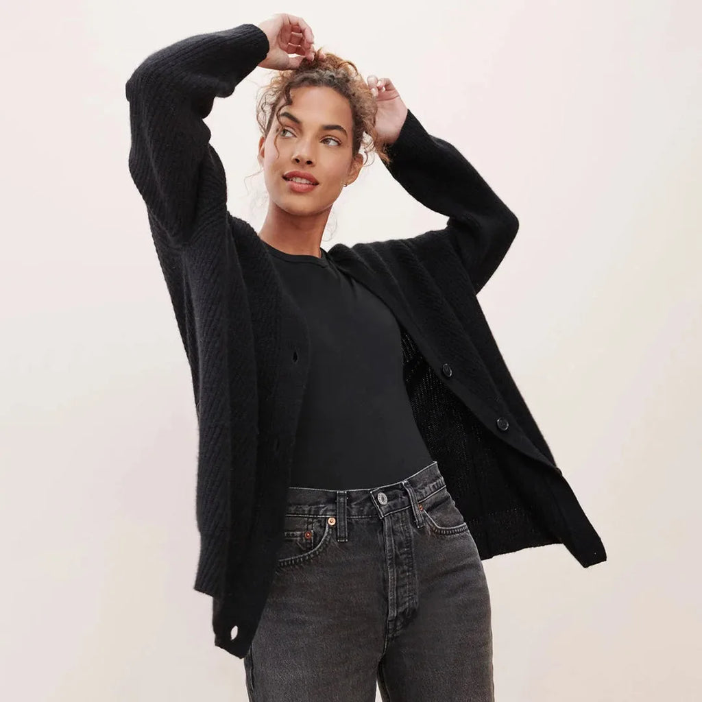 Model lifting her arms while wearing an open black ribbed cardigan over a black top, front view