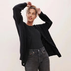 Model lifting her arms while wearing an open black ribbed cardigan over a black top, front view
