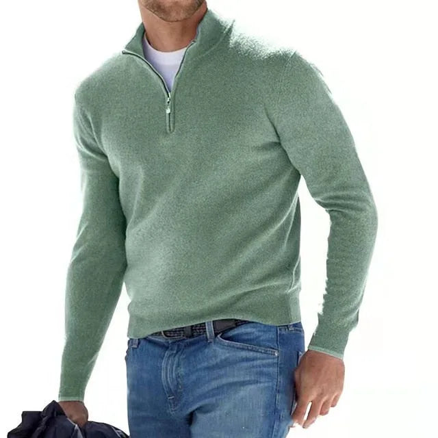 Man wearing a sage green quarter-zip jumper with ribbed collar, paired with jeans and a navy jacket draped over his arm.