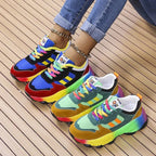 Pair of rainbow sole chunky trainers shown together, one in blue and red, one in green and mustard, on a wooden slat floor