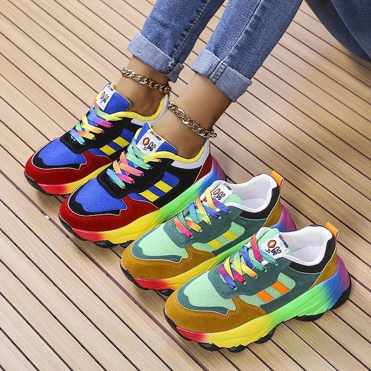 Pair of rainbow sole chunky trainers shown together, one in blue and red, one in green and mustard, on a wooden slat floor