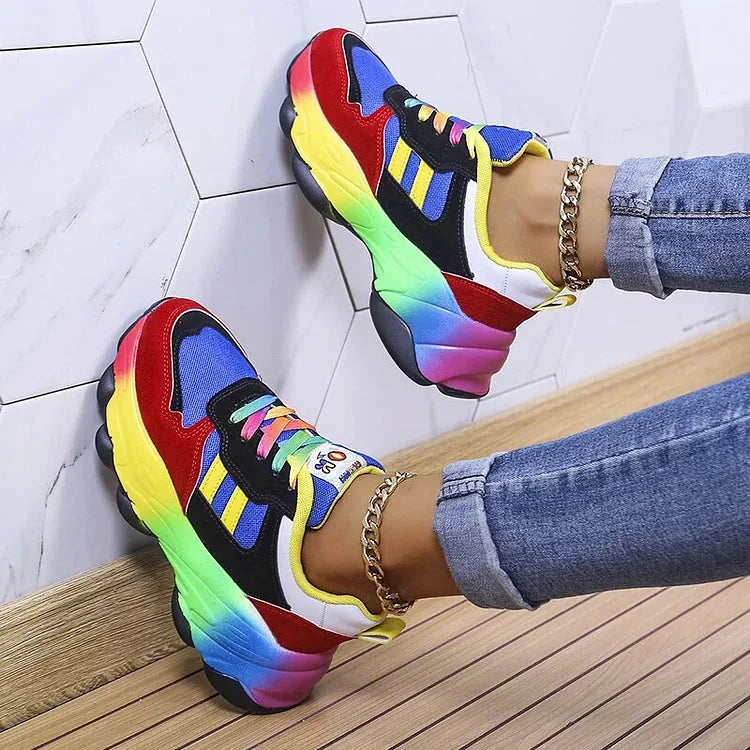 Back view of blue and red chunky trainers against a white hexagon tile wall, showing the gradient rainbow heel