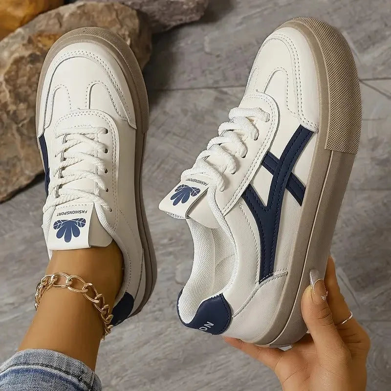 Close-up of white and navy panelled trainers with tan sole, one shoe held by hand showing side stripe detail