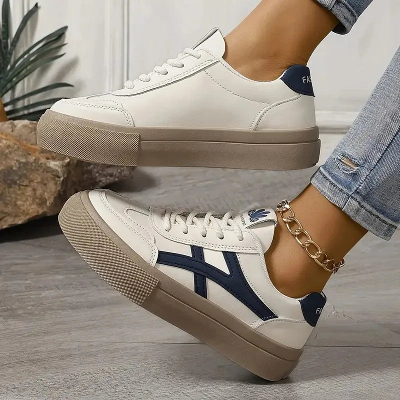 Side view of white leather trainer with navy heel tab and tan sole against a stone background