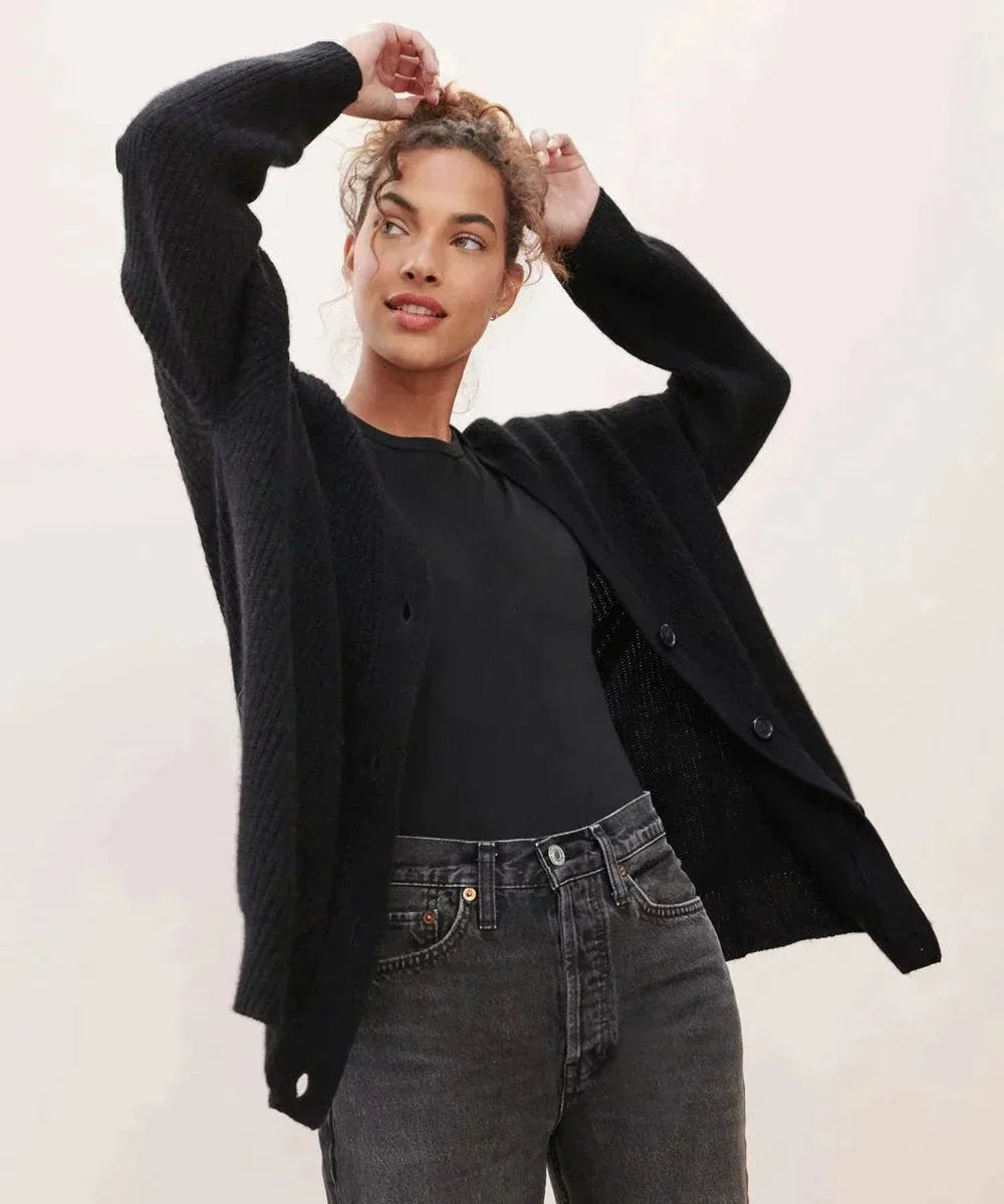 Woman in black ribbed cardigan with arms raised, worn over a black top and dark denim jeans