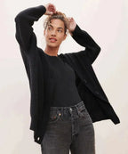 Woman in black ribbed cardigan with arms raised, worn over a black top and dark denim jeans