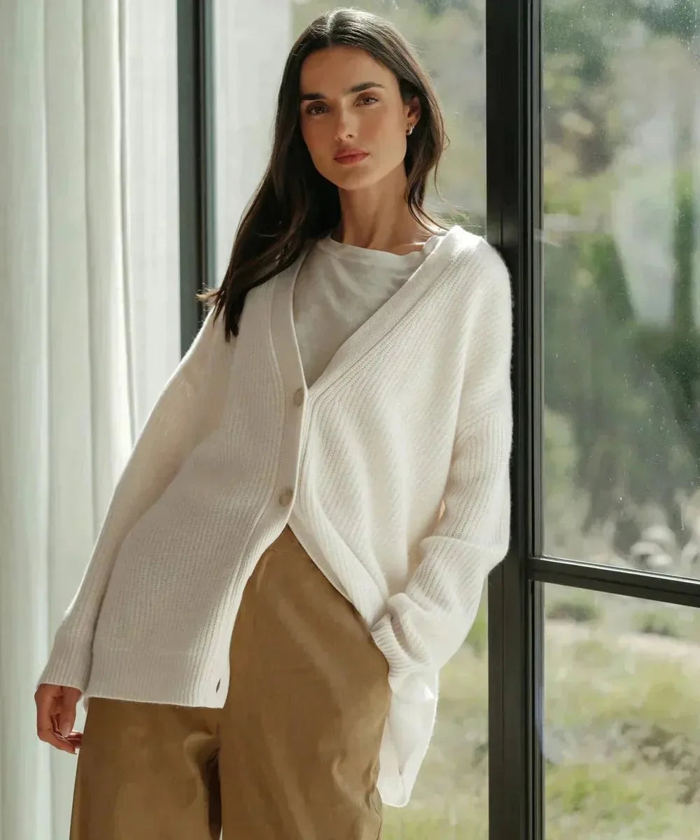 Woman in white ribbed cardigan standing by a window, worn over a white top and tan wide-leg trousers