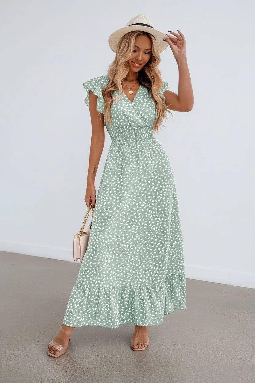 Woman wearing a sage green ditsy print maxi dress with ruffle sleeves and a straw hat, standing against a white wall
