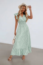 Woman wearing a sage green ditsy print maxi dress with ruffle sleeves and a straw hat, standing against a white wall