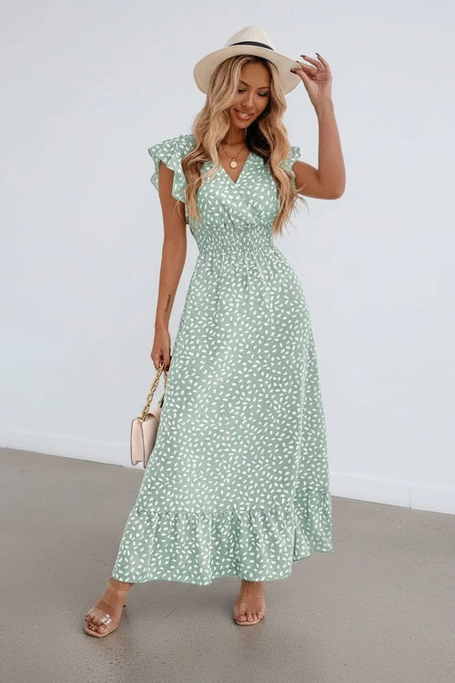 Woman wearing a sage green ditsy print maxi dress with ruffle sleeves and a straw hat, standing against a white wall