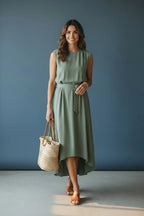 Front view of the sage green midi dress paired with tan slide sandals and a straw tote, standing against a blue backdrop.