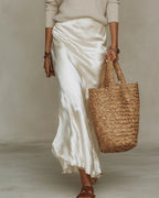 Full-length side view of the ivory satin slip dress falling to ankle length, styled with strappy sandals and a straw tote