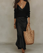 Model walking in a black satin slip skirt paired with a black ribbed V-neck jumper, carrying a woven straw tote
