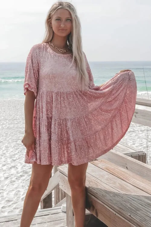 Pink sequin tiered mini dress worn on a beach boardwalk, model holding the skirt out to show the sparkling fabric texture