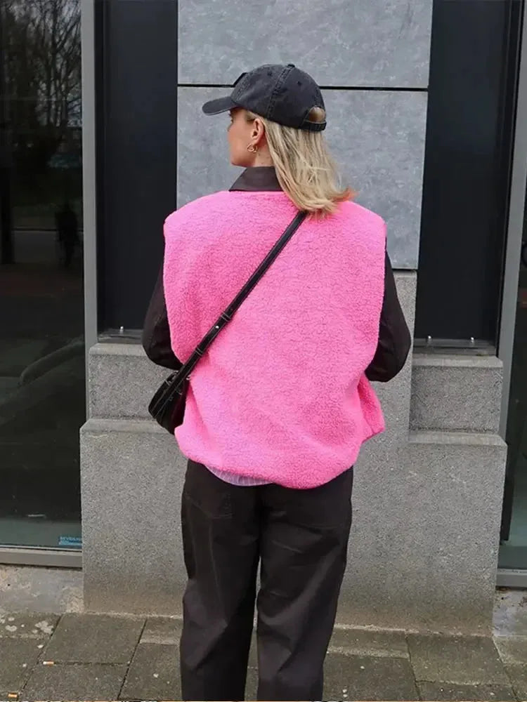 Back view of a bright pink sherpa fleece body warmer worn with a crossbody bag and dark trousers