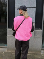 Back view of a bright pink sherpa fleece body warmer worn with a crossbody bag and dark trousers