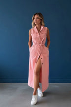 Woman standing in a sleeveless blush pink denim shirt dress with tie waist belt and front button placket, worn with white trainers.
