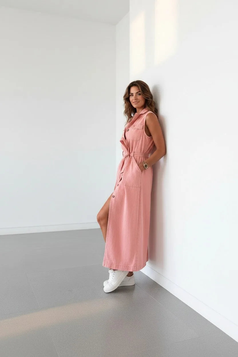 Side profile of the sleeveless denim dress leaning against a wall, highlighting the A-line skirt length and tie belt detail.