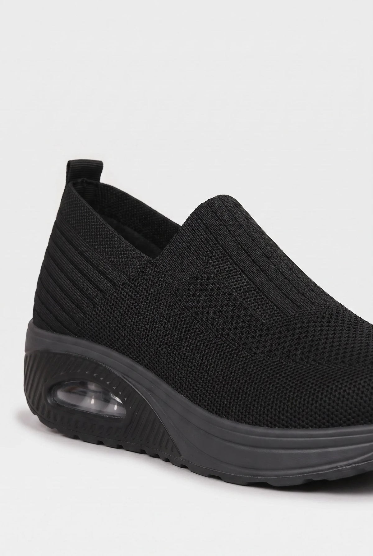 Close-up angled view of black knit slip-on trainer showing ribbed texture and pull tab at the heel