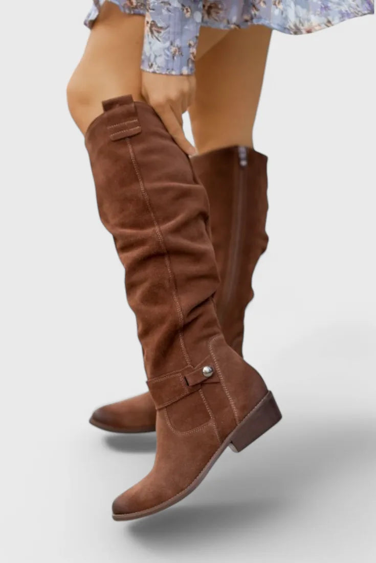 Side view of slouchy brown suede knee-high boot with tonal stitching and buckle tab, worn with a floral dress