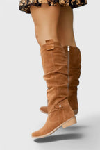 Close-up of tan suede knee-high boot showing slouch folds, buckle tab and stacked block heel