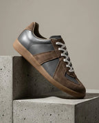 Grey leather trainer with brown suede panels and white laces, gum sole, angled side profile on stone