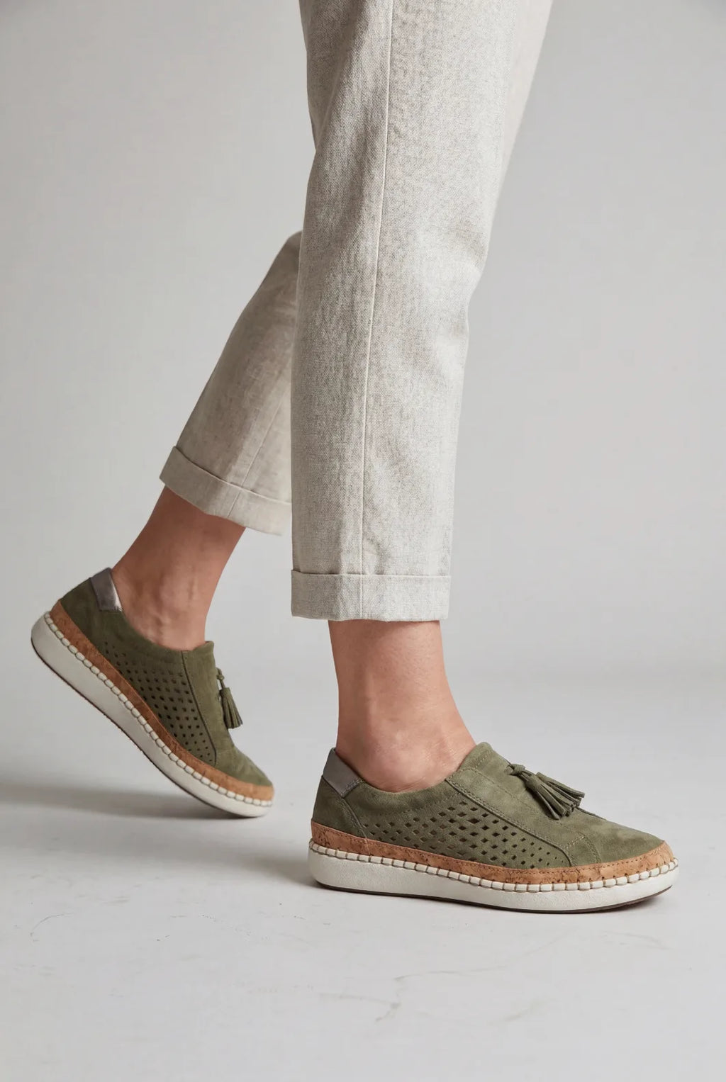 Model walking in cream trousers wearing green suede slip-on trainers with tassel detail and white stitched platform sole