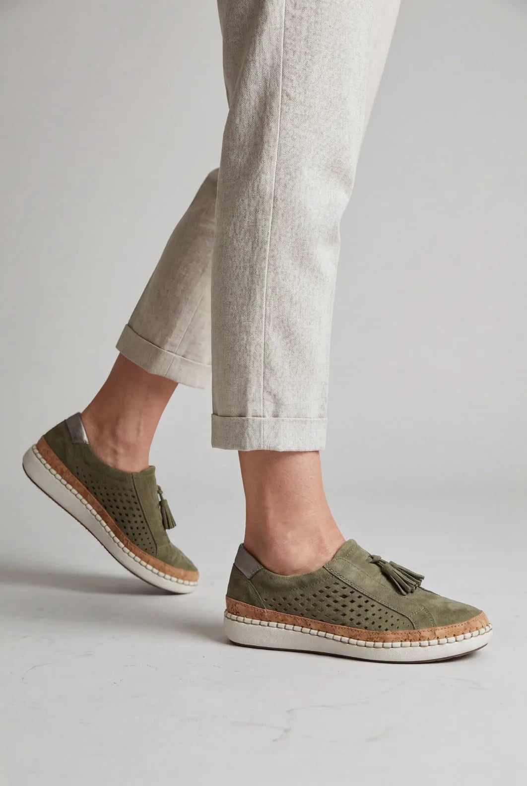 Model walking in cream trousers wearing green suede slip-on trainers with tassel detail and white stitched platform sole