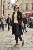 Woman in a khaki wool coat and grey trousers walking outdoors wearing green suede slip-on trainers on a cobbled street