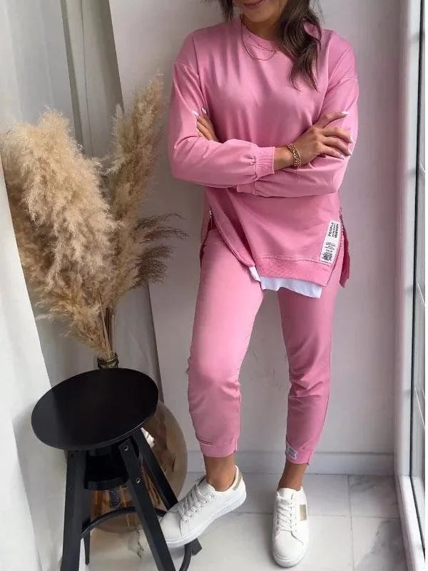 Pink long-line sweatshirt and matching joggers worn with arms crossed, showing the side zip and patch label