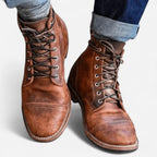 Close-up of both feet wearing tan brown leather lace-up boots with cap toe stitching, paired with rolled denim jeans
