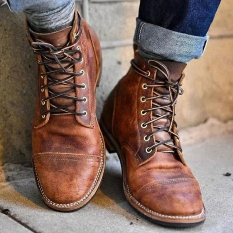 Front view of tan leather lace-up boots with distressed finish and contrast dark eyelets, worn against a stone wall
