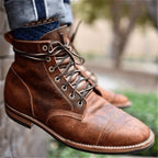 Side profile of a single tan leather boot in mid-air, showing the cap toe seam, lacing detail and stacked heel