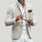 Older man in the white textured jacket buttoned over a white shirt, holding sunglasses, with white trousers