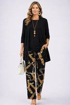 Woman in a black longline open-front cardigan with black round-neck top and gold chain-print trousers, holding a cream handbag
