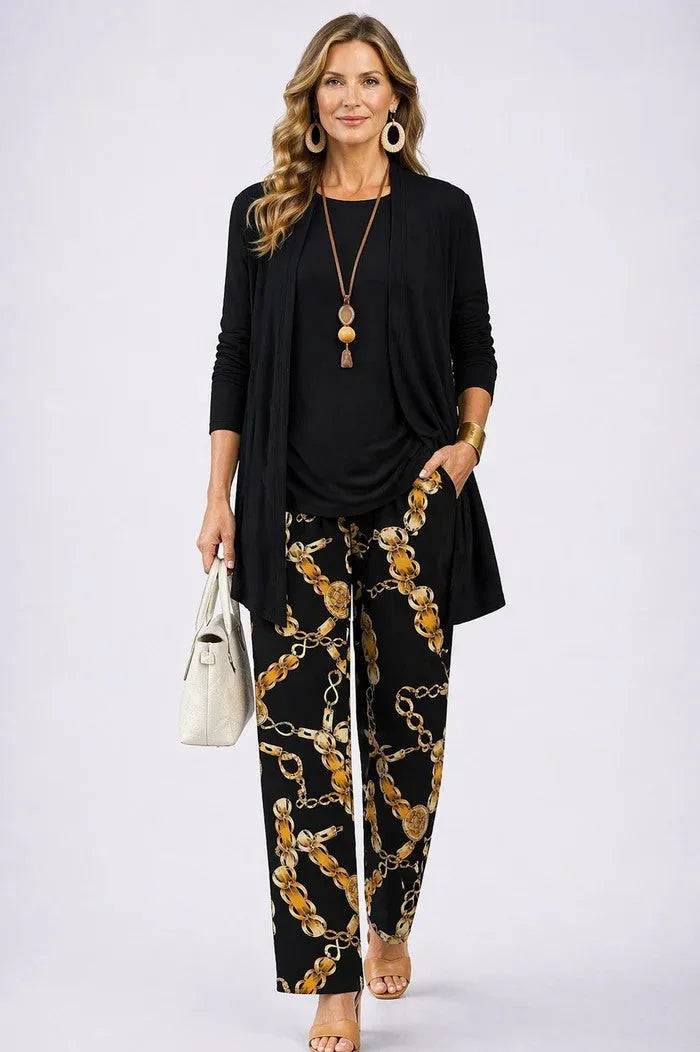 Woman in a black longline open-front cardigan with black round-neck top and gold chain-print trousers, holding a cream handbag