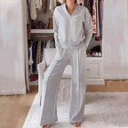 Woman wears a heather grey V-neck jumper and wide-leg trousers with front seam detail, standing in front of an open wardrobe