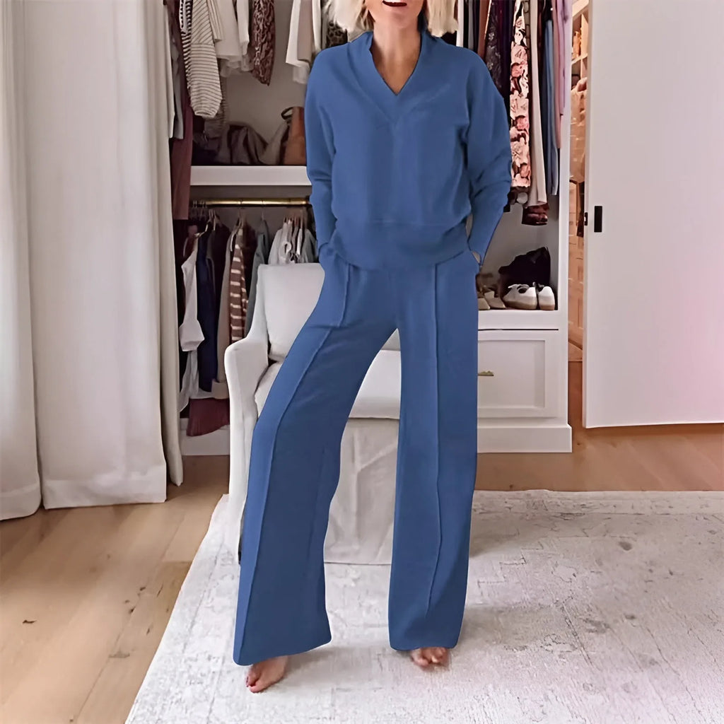 Model in a slate blue V-neck jumper and matching wide-leg trousers standing in a bedroom with an open wardrobe