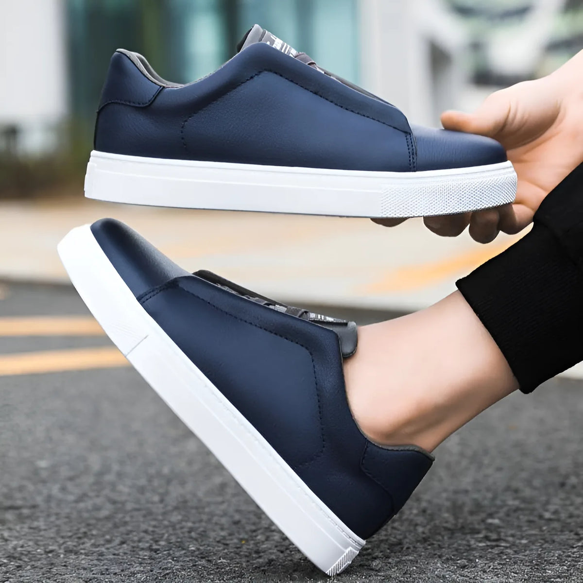 Navy blue leather trainer held in hand above a matching pair worn on foot outdoors
