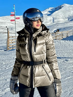 Woman on a snowy ski slope wearing a metallic silver belted puffer ski jacket with black zip trim and matching hood