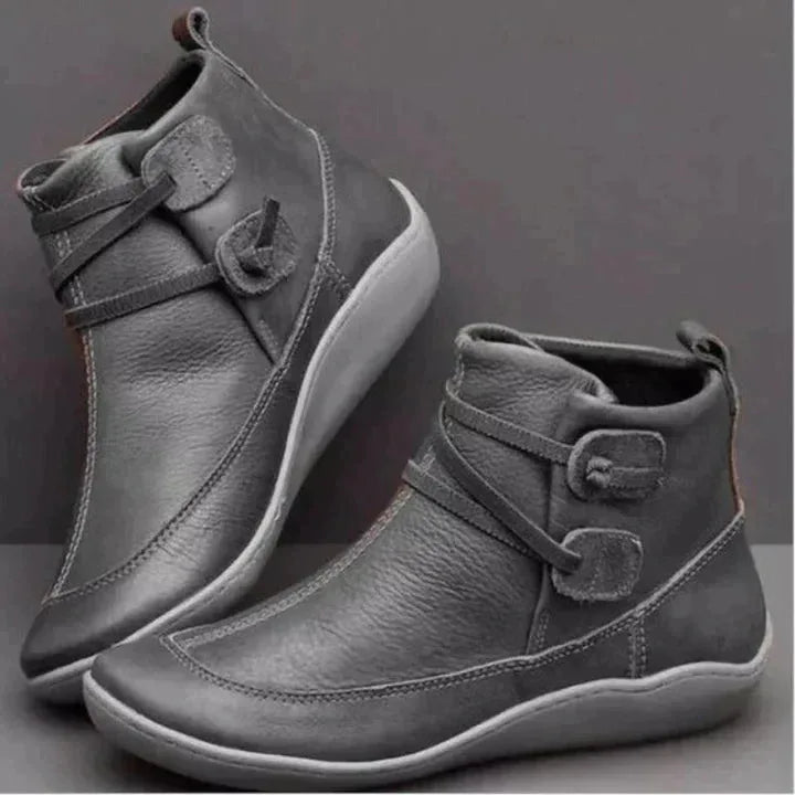Grey leather ankle boots shown from the side with round toe, contrast white sole and criss-cross strap buckle