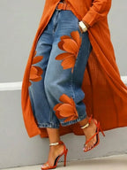 Orange floral print denim trousers worn with a matching orange coat and strappy red heeled sandals with gold ankle strap