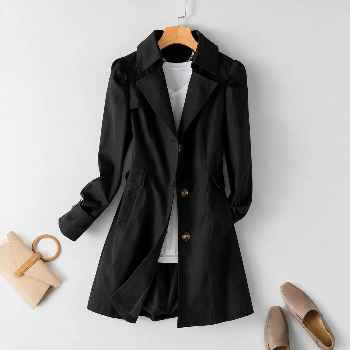 Black trenchcoat with puffed sleeves and flap pockets hanging on a wooden hanger, styled flat lay with loafers and a clutch bag